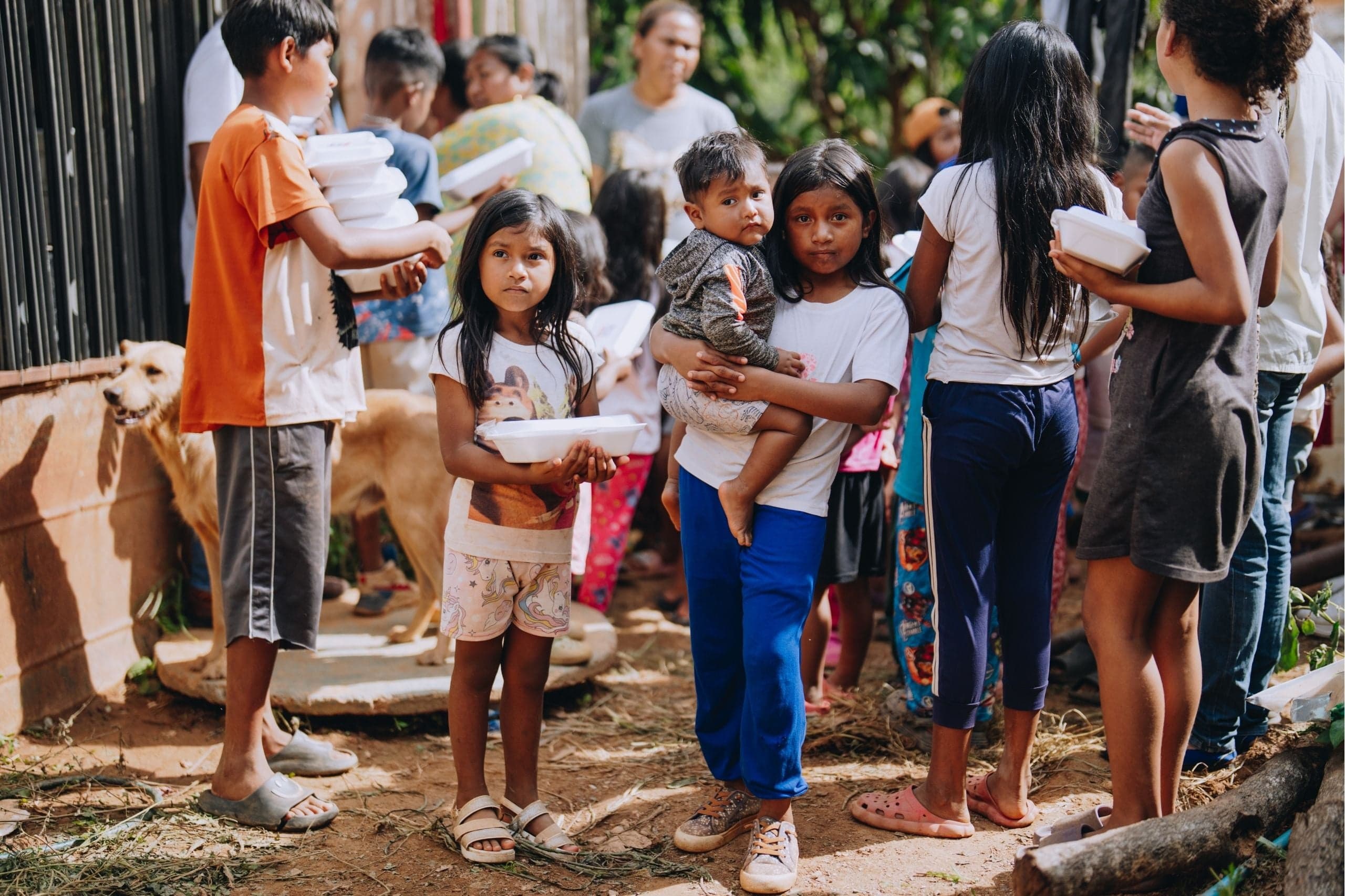 Two girls—one holding a meal and another a baby—look at the camera.