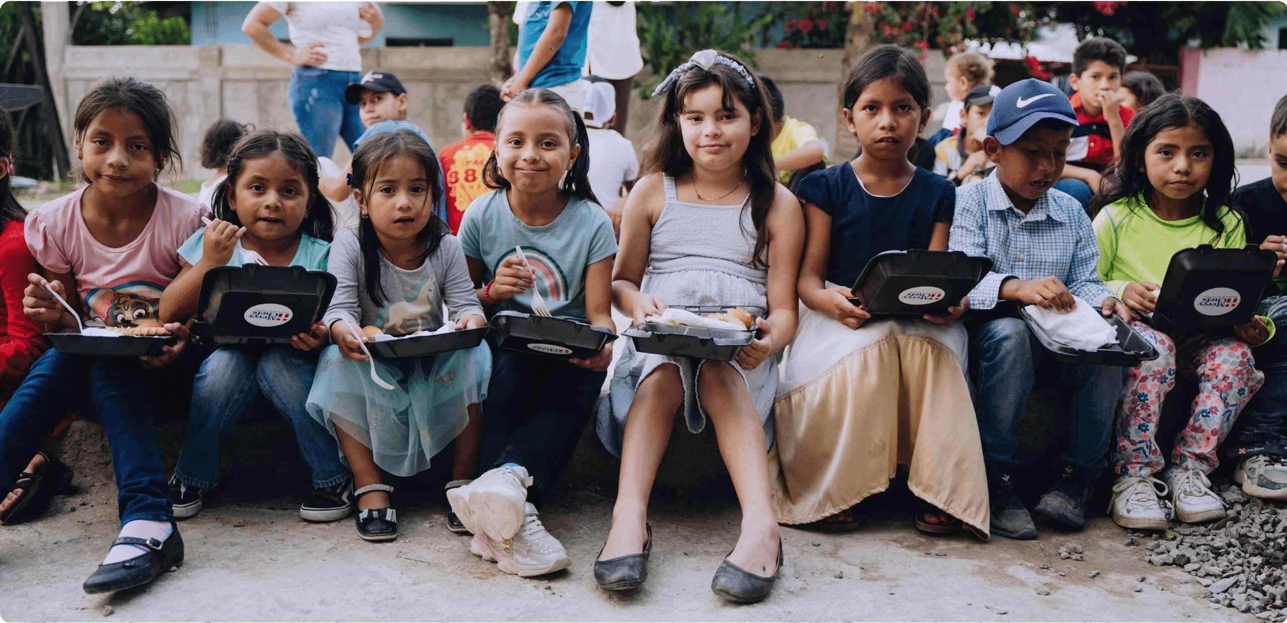 Children in Honduras smile as they eat Mercy Chefs meals siting on the street